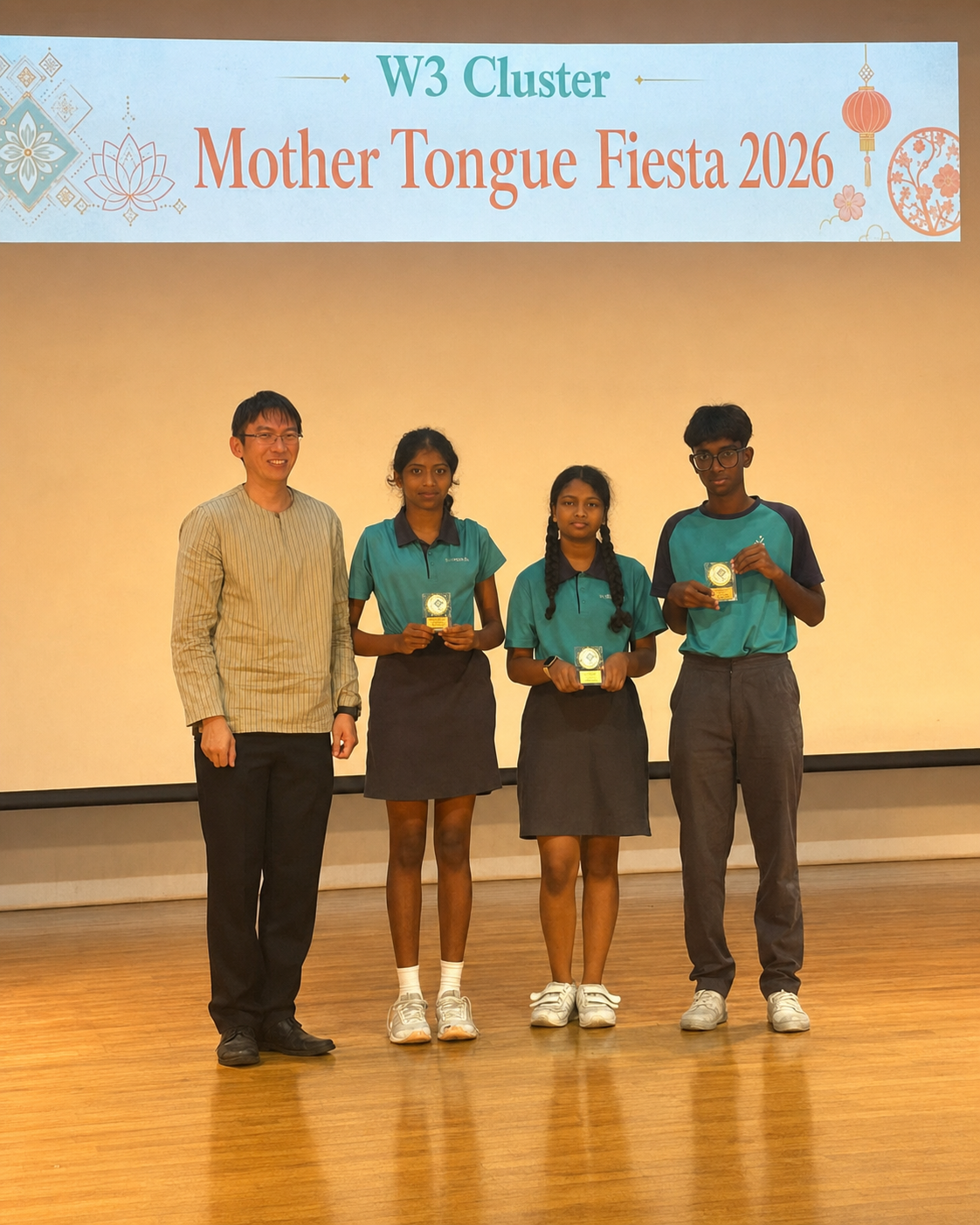 Three students in teal uniforms hold trophies beside a man at the Mother Tongue Fiesta.